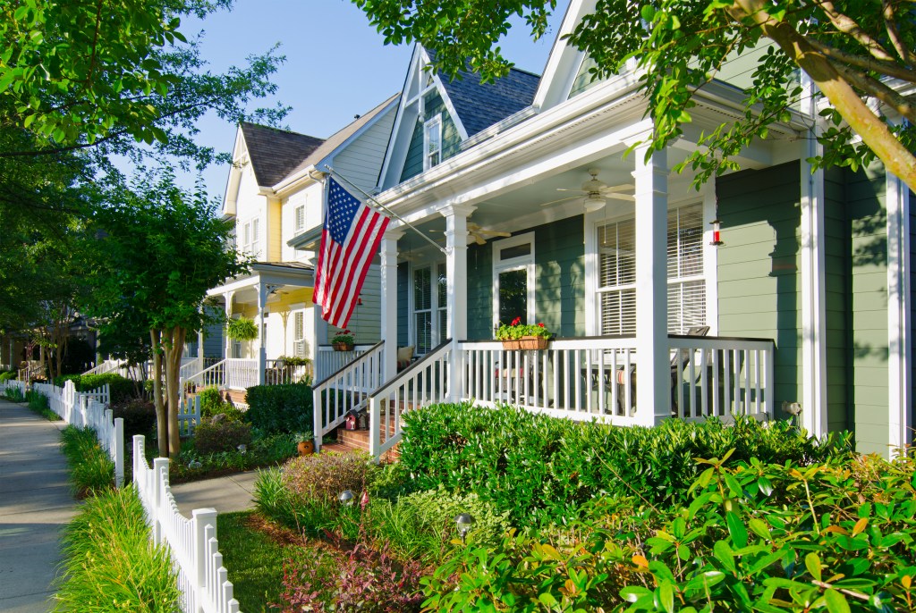 Fort Mill, South Carolina, USA - June 14, 2014: The American Dream is captured in this iconic image of a row of new Victorian-style homes with a white picket fence in the Baxter Village neighborhood development located south of Charlotte, Carolina of the North. Most American homes are now built on smaller lots with sidewalks and tree-lined streets on the outskirts of big cities. An American flag hangs from the front porch to honor an upcoming holiday. Many Americans express their patriotism by flying Old Glory, not just on holidays, but year-round.