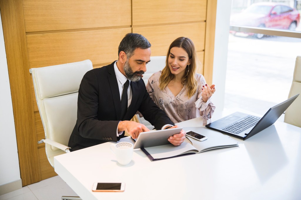 A mature man and young woman, business partners, working in an office, using a digital tablet and laptop respectively